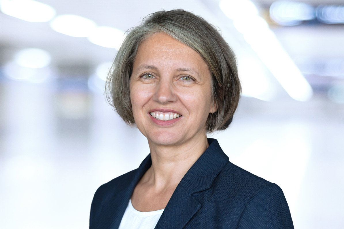 Porträt einer lächelnden Frau mittleren Alters mit kurzen, ergrauten Haaren, dunkelblauem Blazer und weißem Oberteil vor einem unscharfen hellen Hintergrund.