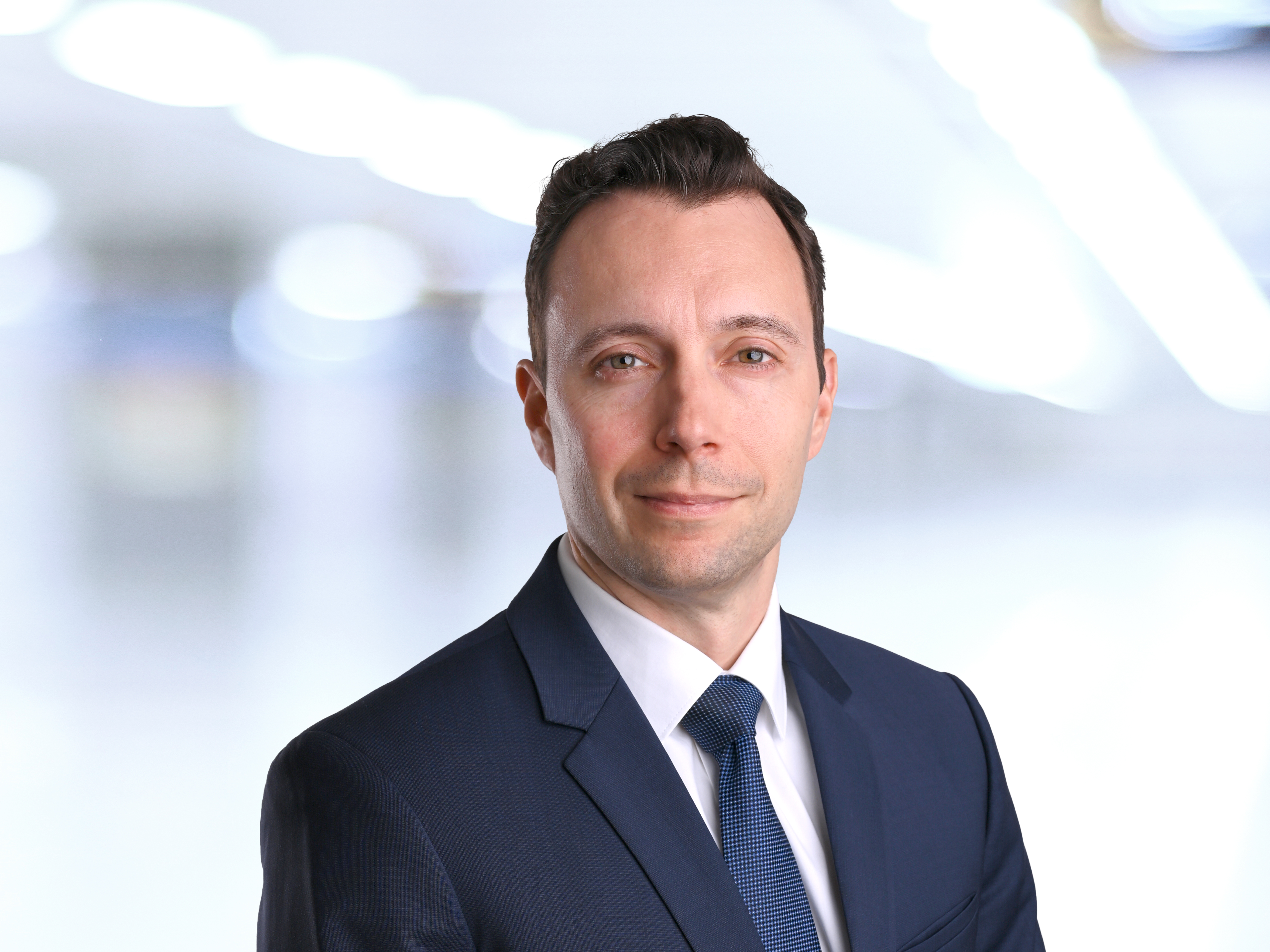 Professionelles Porträtfoto eines Mannes mit kurzen braunen Haaren im dunkelblauen Anzug, weißem Hemd und blauer Krawatte vor einem hellen, verschwommenen Büro-Hintergrund.